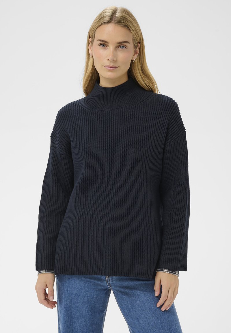 Femme blonde portant un pull col roulé côtelé bleu marine et un jean, se tenant devant un fond blanc uni.