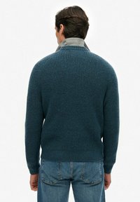 Dunkelblauer Strickpullover mit gerippter Textur, Rundhalsausschnitt und langen Ärmeln, kombiniert mit einem grauen Hemd mit Kragen darunter.