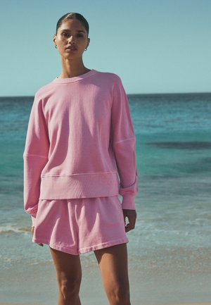 Růžová bavlněná mikina s dlouhými rukávy a volným střihem, doplněná o odpovídající šortky. Jemná textura, neformální design bez viditelných vzorů.