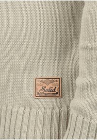 Gebreide trui in lichtbeige met een geribbelde textuur. Voorzien van een bruine leren patch met het label "Solid United Traditions" en decoratieve elementen.