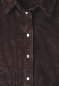 Chemise en velours côtelé brun foncé avec des rainures verticales et des boutons-pression métalliques sur le devant sous un col pointu.