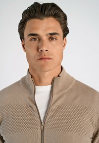 Beżowy sweter na zamek z teksturowanym wzorem w romby na klatce piersiowej, ściągaczowy kołnierz i biały podkoszulek. Gładki materiał, casualowy design.