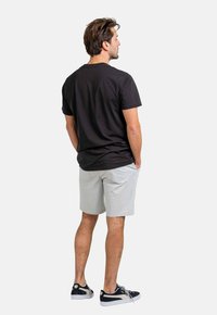Camiseta de algodón negra de manga corta, combinada con pantalones cortos gris claro. Lleva zapatillas negras con suelas blancas. Fondo neutro.