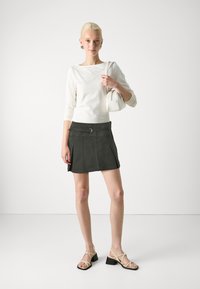 Haut à manches longues blanc, mini-jupe plissée verte foncée avec ceinture, et escarpins beiges à lanières. Le mannequin tient un petit sac à main blanc.