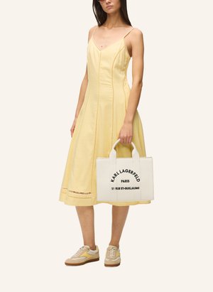 Naine, kes kannab kollast varrukateta keskmise pikkusega kleiti ja beeže tenniseid, hoides valget Karl Lagerfeldi käekotti musta tekstiga.