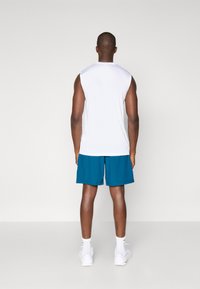 Ärmelloses weißes Sportoberteil mit Rundhalsausschnitt, kombiniert mit blauen Shorts. Das Modell trägt weiße Sneaker und knöchellange Socken.