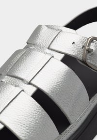 Ασημί σανδάλι από υφή δέρματος με φαρδιές λουρίδες και ρυθμιζόμενο μεταλλικό αγκράφα σε μαύρη σόλα, απεικονίζεται σε κοντινό πλάνο.