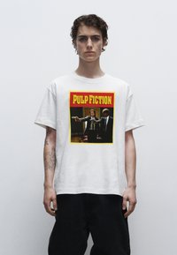 Bílý bavlněný tričko s barevným potiskem "PULP FICTION" zobrazujícím dvě postavy mířící zbraněmi, orámované žlutou a červenou.