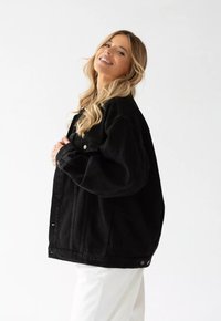 Czarna kurtka dżinsowa o luźnym kroju, z dużymi kieszeniami, z przodu zapinana na guziki, wykonana z lekko teksturowanej tkaniny. Noszona na białych spodniach.