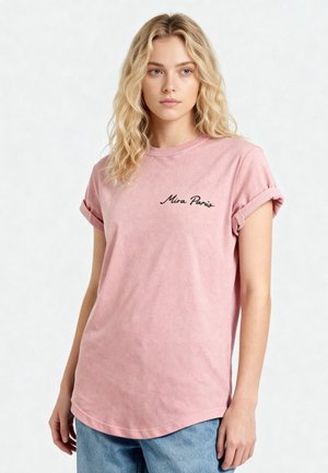 Jonge vrouw met golvend blond haar, die een roze t-shirt met opgerolde mouwen en de tekst "Mira Paris" draagt, en een blauwe spijkerbroek, staand tegen een witte achtergrond.