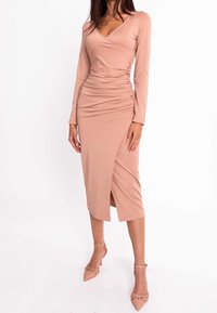 Femme portant une robe midi beige à manches longues, froncée et portefeuille, avec des talons à brides à la cheville et bout pointu sur fond blanc.