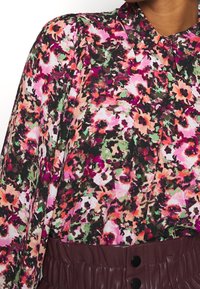 Blouse florale avec des nuances de rose, de vert et de noir ; tissu léger ; patte de boutonnage ; épaules légèrement bouffantes ; associée à une ceinture bordeaux.