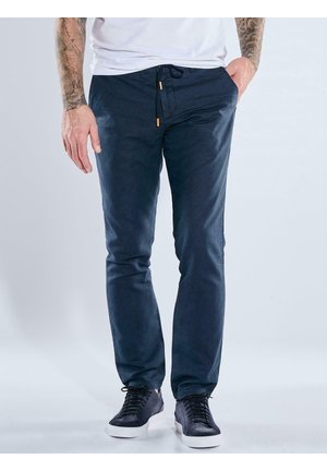 Man wearing navy blue slim-fit pants with drawstring and black sneakers, standing with one hand in pocket against plain background.