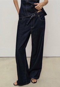 Pantalones anchos de mezclilla azul oscuro con cintura alta, que presentan un cinturón y costuras de acento. Textura suave con un ajuste relajado y bolsillos.