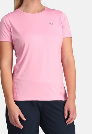 Kvinde iført lyserød kortærmet sportsbluse med lille logo på brystet og mørkeblå bukser, stående mod en hvid baggrund.