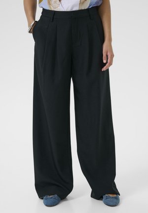 Melnas plašas bikses ar krokojumu, izgatavotas no gludas auduma. Ir sānu kabatas un vaļēja piegriezuma, saskaņotas ar zilām plakanām kurpēm.