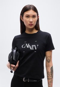 Μαύρο βαμβακερό t-shirt με λευκό σχέδιο «Calvin Klein New York». Συνδυασμένο με μικρή μαύρη δερμάτινη τσάντα και μαύρη ζώνη.