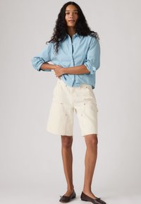 Γυναίκα με μακριά σγουρά μαλλιά, που φοράει ανοιχτό μπλε πουκάμισο με κουμπιά, κρεμ βερμούδα μέχρι τα γόνατα με τσέπες και σκούρα καφέ φλατ παπούτσια, στέκεται μπροστά σε απλό φόντο.