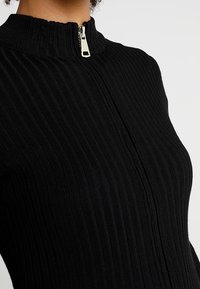 Robe noire en maille côtelée avec fermeture éclair au niveau du cou. Le tissu présente un motif texturé, créant des lignes verticales sur le vêtement.