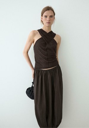 Femme portant un haut sans manches marron foncé à bretelles croisées et une jupe ample marron foncé taille haute, tenant un sac à main rond noir tressé.