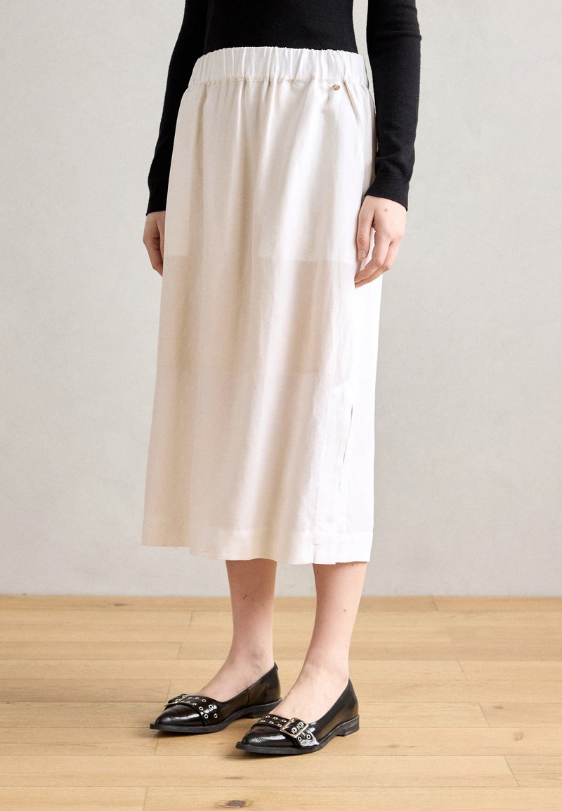 Jupe midi blanche avec une taille élastique, fentes latérales et une texture lisse, accompagnée de chaussures plates noires avec des boucles décoratives.