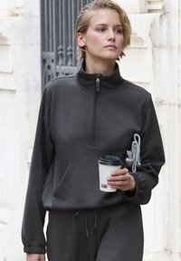Felpa grigia scura con zip, colletto alto, tasca frontale e tessuto texturizzato. Tiene un bicchiere di caffè usa e getta, con dettagli in corda grigia.