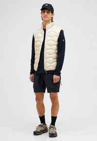 Gepolsterte cremefarbene Weste mit wellenförmiger Stepptextur, schwarzes langärmliges Hemd darunter, passende schwarze Shorts und braune Wanderschuhe.