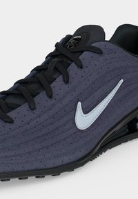 Σκούρο μπλε αθλητικό παπούτσι Nike με μαύρα κορδόνια, διάτρητο άνω μέρος, λευκό λογότυπο Swoosh και μαλακή σόλα σε απλό φόντο.