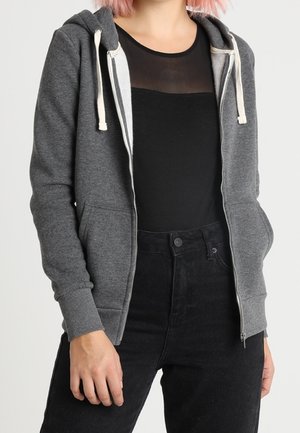 Personne portant un sweat à capuche gris zippé par-dessus un top noir transparent et un pantalon noir taille haute, debout avec une main détendue le long du corps.