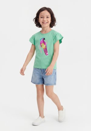 Pige smilende og gående, iført en grøn T-shirt med et farverigt papegøje pailletdesign, denimshorts og hvide slip-on sko.