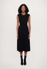 Vestido preto midi com corpete canelado e saia fluida. Design sem mangas, feito de tecido suave. Usado com meias até ao tornozelo e sapatos de salto agudo.