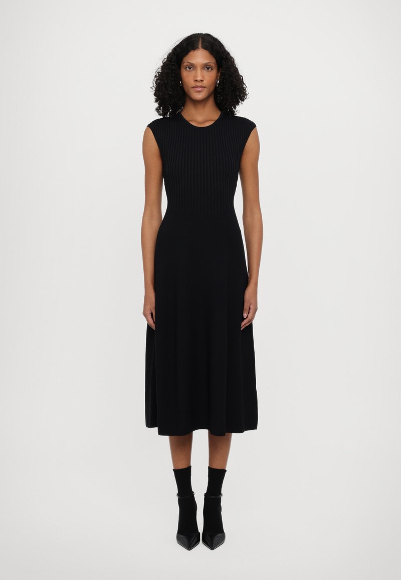 Vestido preto midi com corpete canelado e saia fluida. Design sem mangas, feito de tecido suave. Usado com meias até ao tornozelo e sapatos de salto agudo.