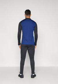 Μπλε αθλητική μπλούζα με μαύρα μανίκια, συνδυασμένη με μαύρες φόρμες και μαύρα αθλητικά παπούτσια, προβαλλόμενη από πίσω.