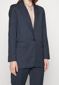 Femme portant un costume à fines rayures bleu marine avec un blazer boutonné d'un seul bouton et un pantalon assorti, accessoirisée d'un collier chaîne argenté épais avec pendentif.