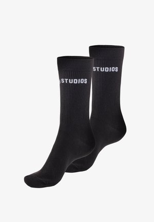 Schwarze gerippte Socken mit weißem "STUDIOS"-Schriftzug oben. Hergestellt aus weichem Material mit Standard-Länge bis zur Wade und nahtlosem Zehendesign.