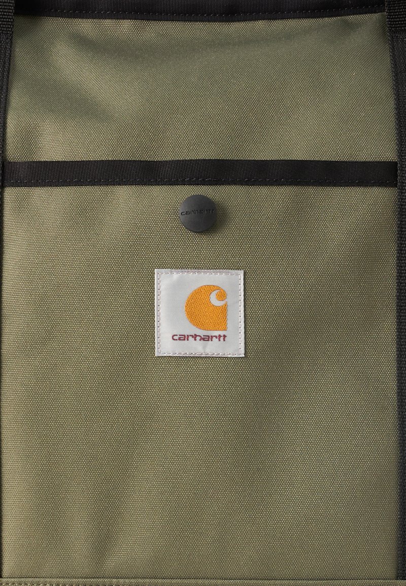 Panel de tela verde oliva claro con ribete negro, un pequeño botón a presión negro encima de una etiqueta cuadrada blanca que muestra un logotipo naranja de Carhartt.