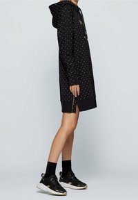 Černé šaty s kapucí a bílými puntíky, vybavené přední kapsou, stahovací kapucí a bočními zipy. Navrženo v uvolněném střihu.