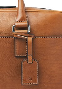Brown leather bag with handle, stitched detailing, metal buckle, and attached rectangular leather tag with embossed logo.