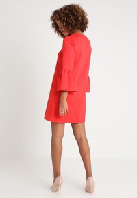 Rotes Kleid mit Glockenärmeln aus glattem Stoff, knielang und mit geradem Schnitt. Kombiniert mit nudefarbenen High Heels.
