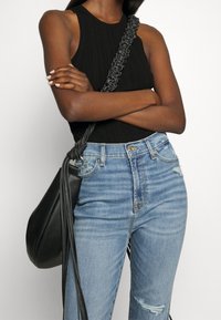 Top de tirantes negro acanalado combinado con jeans de cintura alta desgastados de color azul claro. Un bolso de cuero negro con una correa trenzada reposa de lado.