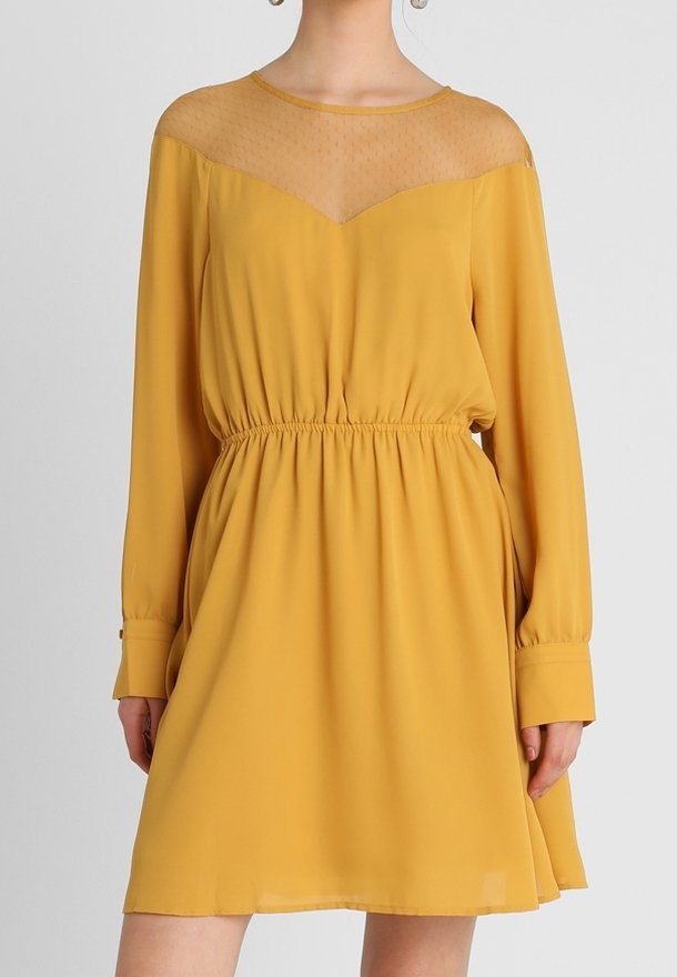 Robe jaune moutarde à manches longues avec taille élastique, col transparent à pois et poignets boutonnés portée par une personne debout.