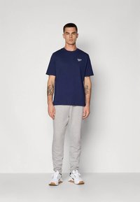 Vīrietis stāv pie baltas sienas, valkājot tumši zilu Reebok T-kreklu, gaiši pelēkas sporta bikses un baltas sporta kurpes ar gumijas zolēm.