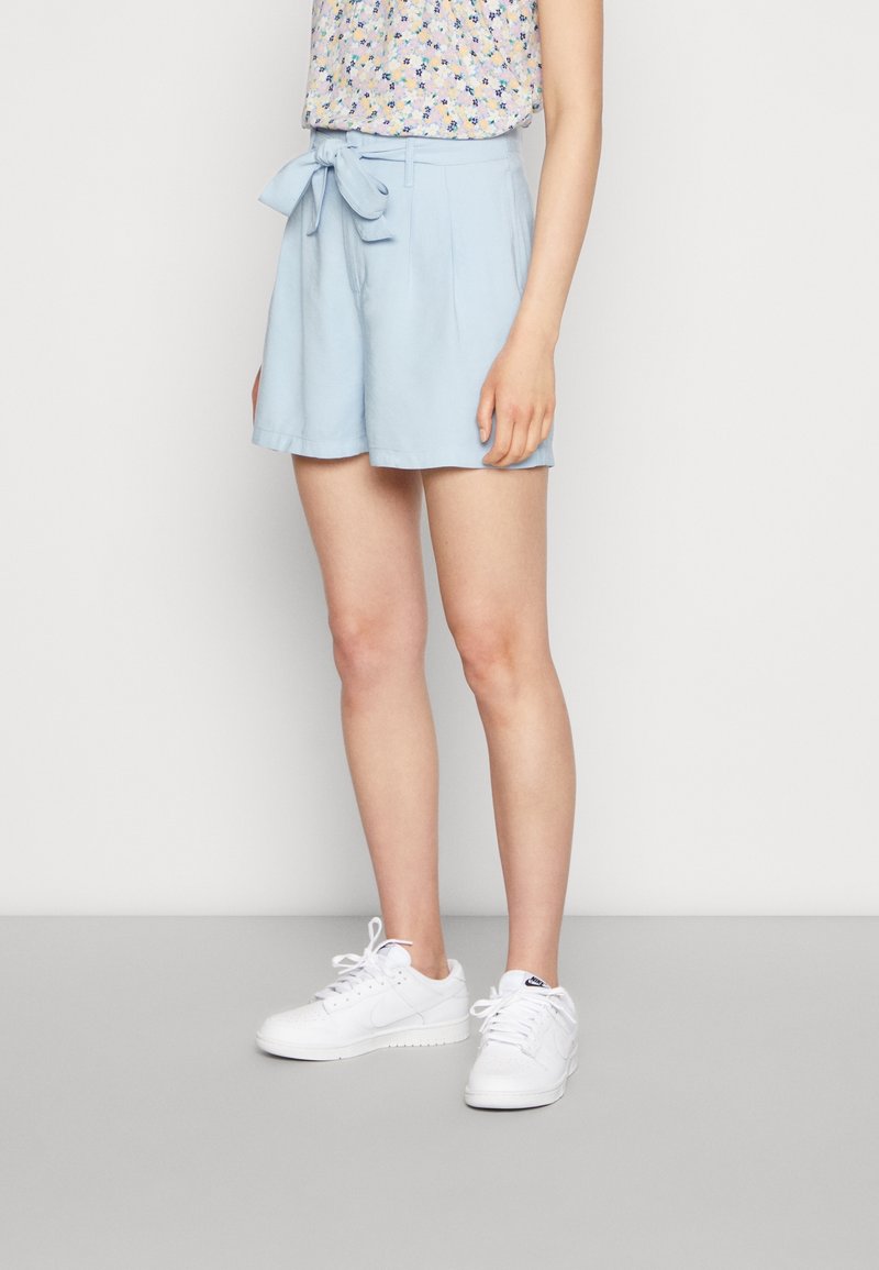 Svetle modré šortky s vysokým pasem, s opaskem na zavázání, sklady a volným střihem, ve spojení s bílými teniskami a květinovým topem.