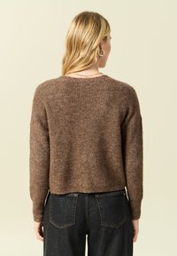 Maglione a coste marrone con design corto e maniche lunghe, indossato sopra jeans di denim scuri, visto di schiena su uno sfondo neutro.