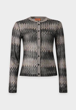 Un mannequin porte un bob noir, un cardigan à motifs, un chemisier blanc, un pantalon marron à jambes larges et des mocassins couleur crème sur un fond uni.