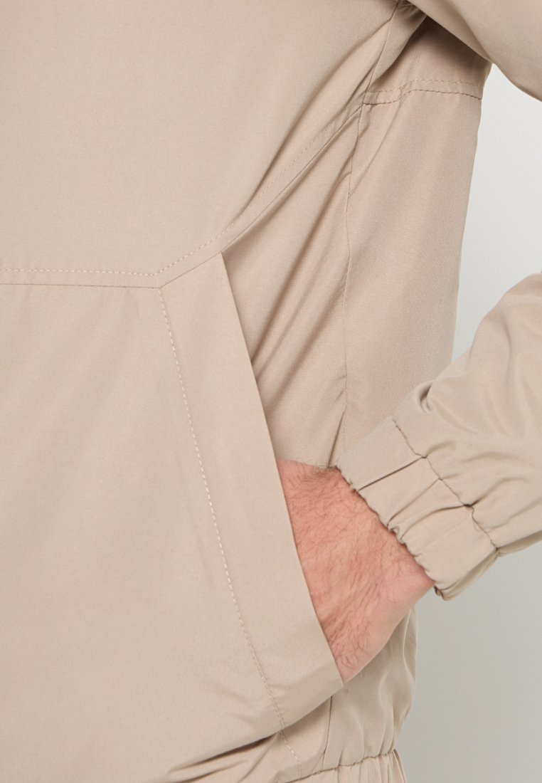 Chaqueta beige con un tejido suave y ligero, puños elásticos y bolsillo lateral, que presenta un diseño minimalista y costuras limpias.