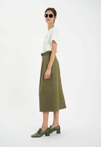 T-shirt blanc avec texte brodé, associé à une jupe midi olive et des chaussures à talons carrés assorties. Lunettes de soleil et coupe décontractée.