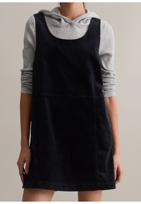 Femme portant une robe salopette en velours côtelé noir par-dessus un sweat à capuche gris clair côtelé avec les manches retroussées, sur un fond clair uni.