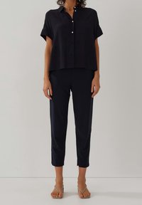 Camicetta nera a maniche corte con dettagli di bottoni, abbinata a pantaloni neri slim-fit e sandali con cinturino in tonalità neutra, su uno sfondo chiaro.