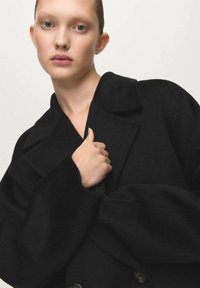 Czarny płaszcz z mieszanki wełny, z klapowym kołnierzem, luźnym krojem i szerokimi rękawami. Charakteryzuje się gładką teksturą i subtelnymi guzikami jako akcentami.